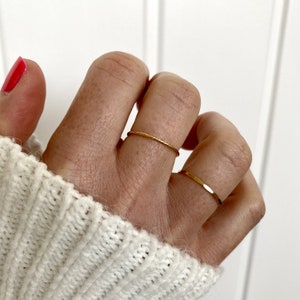 May include: Two delicate gold rings are shown on a hand with red nail polish. One ring has a textured, twisted design, the other is a simple, flat band. The rings are set against a white background.