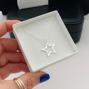 May include: A silver-toned necklace featuring a hollow star-shaped pendant suspended from a delicate chain. The jewelry is presented inside a square white cardboard box, which is nestled within a larger black container.