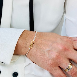 May include: A silver bangle bracelet with a gold infinity symbol charm. The bracelet is on a person's wrist.