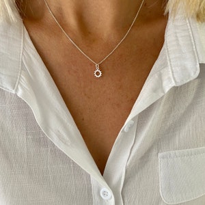 May include: A silver necklace with a small sun-shaped pendant. The pendant has a detailed, open design. The necklace is worn against a white collared shirt.
