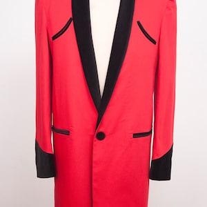 May include: A long, red blazer with black velvet lapels, pocket trim, and sleeve cuffs. The blazer has a single black button closure and is displayed on a mannequin. A classic formal wear garment.