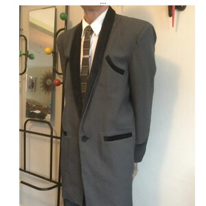 May include: A grey blazer with black lapels and pocket trim. The blazer has a single-button closure and is worn with a white collared shirt and a patterned tie. The suit is a classic style.