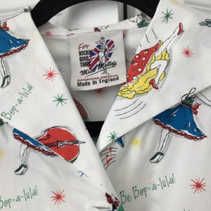 以下が含まれることがあります： ダンスをする人物、楽器、そして「Bop-a-lula!」や「Rock 'n' Roll」という文字がカラフルにプリントされた白い襟付きシャツ。内側のラベルには「For Rockin Good Times Miss Molly's Doghouse Vintage Made In England.」と記載されています。