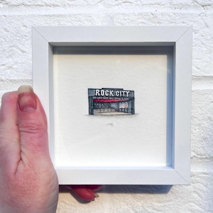 May include: A white framed miniature drawing of a building with the text "ROCK-CITY" above the entrance. The building has a red awning and a grey facade.