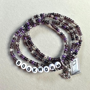 May include: A beaded wrap bracelet with purple, clear, and black beads. The bracelet spells out "BOOKWORM" with white letter beads. A silver charm of a book and a worm is attached.