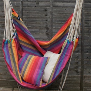May include: A colorful striped hammock chair hangs from a wooden bar. The hammock is made of a soft, woven fabric and has a white pillow inside. The chair is suspended in front of a wooden fence.