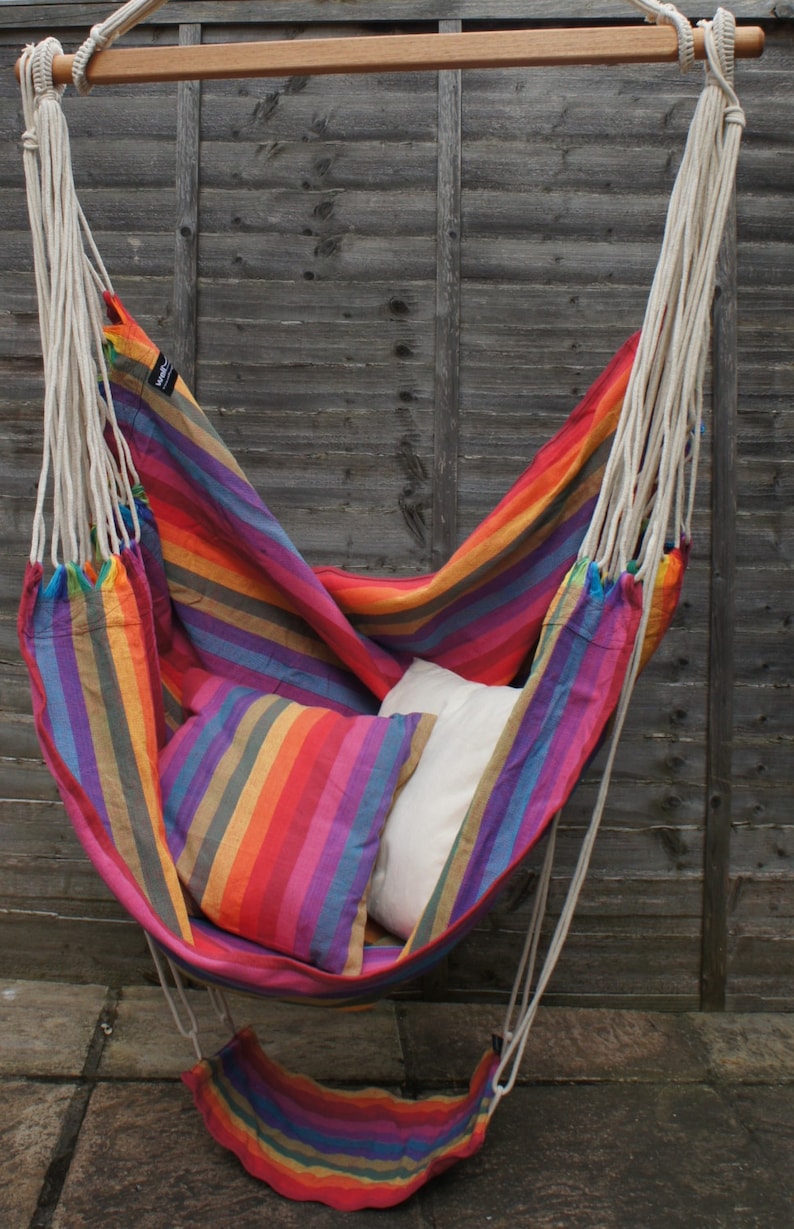 May include: A colorful striped hammock chair hangs from a wooden bar. The hammock is made of a soft, woven fabric and has a white pillow inside. The chair is suspended in front of a wooden fence.