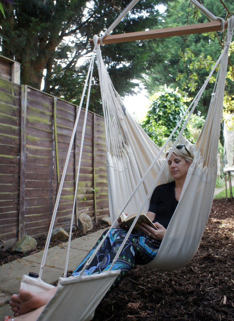 May include: A person relaxing in a white hammock chair, reading a book. The hammock is suspended from a wooden beam and is hanging between two trees. The person is wearing a black shirt and colorful pants.