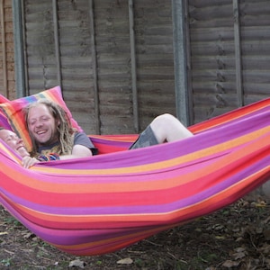 May include: Two people relaxing in a colorful striped hammock with a pillow. The hammock is suspended between two wooden posts.