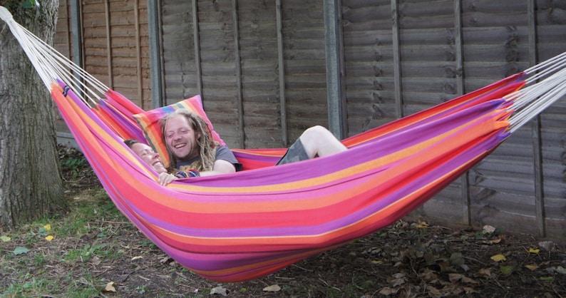 May include: Two people relaxing in a colorful striped hammock with a pillow. The hammock is suspended between two wooden posts.