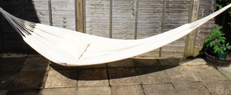 May include: A white hammock hanging between two wooden posts. The hammock is made of a soft, woven fabric and is perfect for relaxing outdoors.