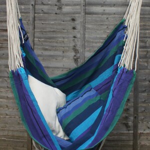 May include: A blue and green striped hammock chair with white ropes hanging from a wooden bar. The hammock is filled with two white pillows.