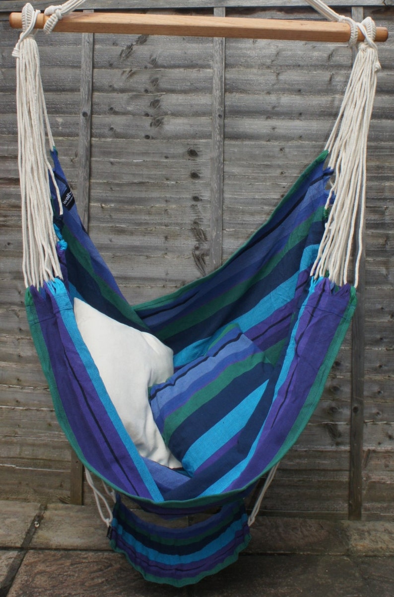 May include: A blue and green striped hammock chair with white ropes hanging from a wooden bar. The hammock is filled with two white pillows.