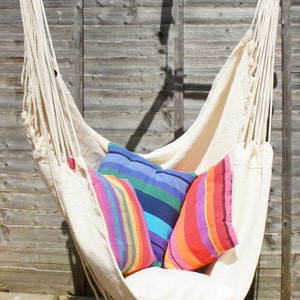 May include: A white hammock chair with three colorful striped pillows. The hammock is hanging from a wooden bar and is suspended in front of a wooden fence.