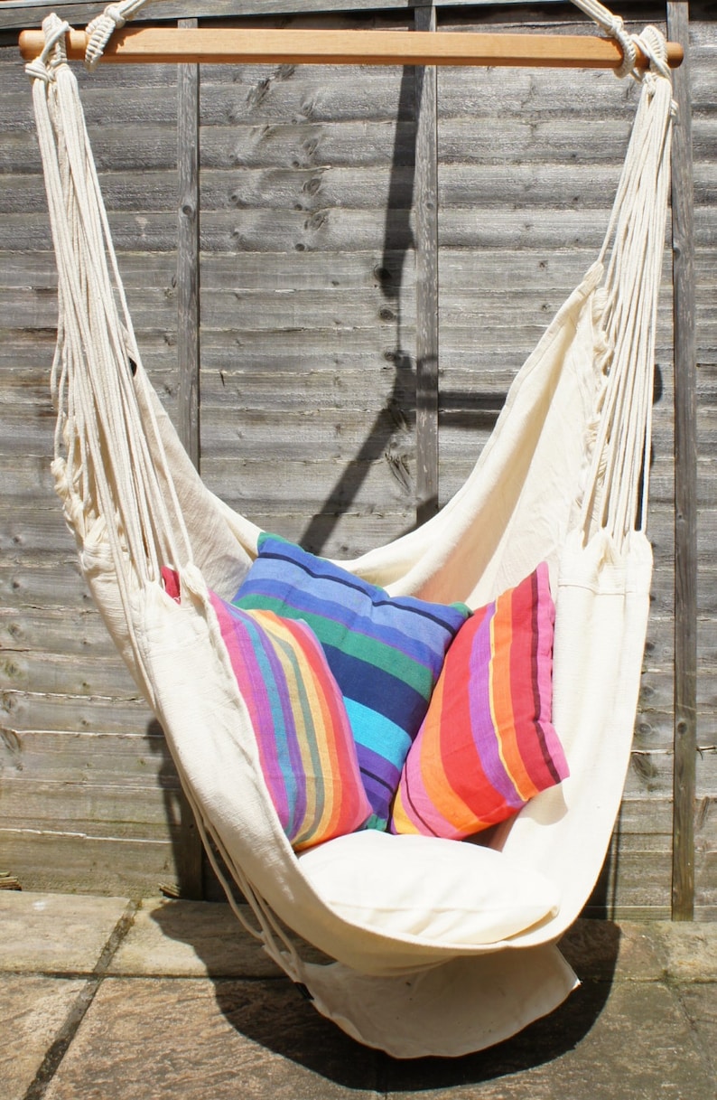 May include: A white hammock chair with three colorful striped pillows. The hammock is hanging from a wooden bar and is suspended in front of a wooden fence.