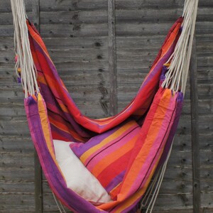 May include: A colorful striped hammock with a white pillow hanging from a wooden beam against a gray wooden fence.