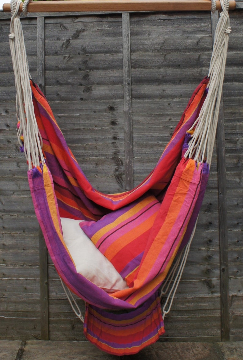 May include: A colorful striped hammock with a white pillow hanging from a wooden beam against a gray wooden fence.