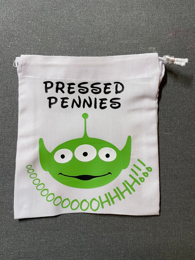 Puede incluir: Bolsa blanca con cord&oacute;n con las palabras "PRESSED PENNIES" en negro. Una cara de alien&iacute;gena verde con tres ojos y la frase "OOOOOOOOHHH!!!" en verde est&aacute; impresa en la parte delantera. La bolsa es de tela.