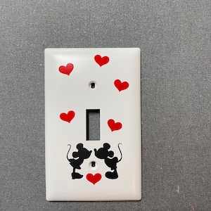 May include: White light switch cover featuring red hearts and silhouettes of Mickey and Minnie Mouse. The black silhouettes are facing each other, with a heart between them. Additional red hearts are scattered around the cover.