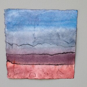May include: A watercolour painting with a colour gradient. The top is blue, fading to grey, then purple, and finally red at the bottom. The paper edges are rough and uneven.