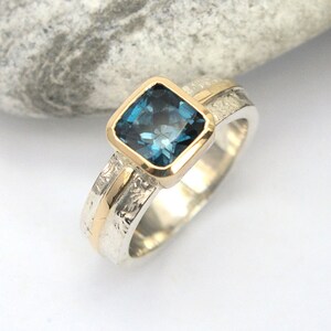 May include: A silver and gold ring with a square blue topaz gemstone. The ring has a textured band.