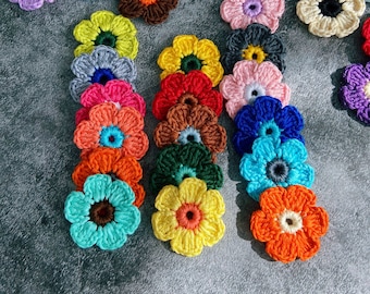 Pode incluir: Uma coleção de flores de crochê coloridas em vários tons de azul, verde, vermelho, amarelo, laranja, rosa, roxo e marrom. As flores estão dispostas em um padrão decorativo sobre uma superfície cinza.
