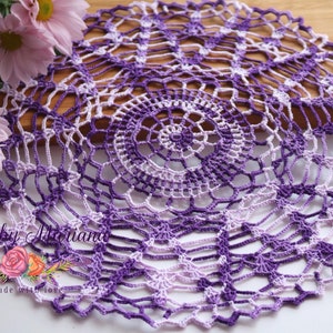 May include: A purple and white crocheted doily with a delicate, lacy design. The doily is handmade and features a gradient of color from light purple to dark purple.