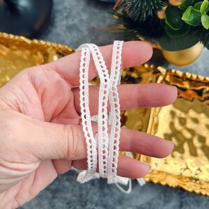 Puede incluir: Primer plano de un ribete de encaje blanco con borde festoneado. El delicado adorno se sostiene en una mano, mostrando su intrincado diseño. El fondo incluye una bandeja dorada y decoraciones navideñas.
