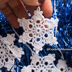 May include: White crocheted snowflakes hanging on a blue tinsel garland. The snowflakes are delicate and intricate, with a small hole in the centre of each one. The text "@CrochetybyMariana" is visible in the bottom right corner of the image.