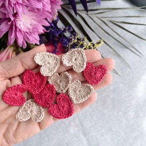 May include: A collection of small, crocheted hearts in shades of red and beige are held in a hand. The hearts are arranged in a scattered pattern. The background features pink flowers and greenery.