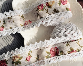 May include: A white lace trim with a floral pattern on a beige fabric ribbon. The ribbon has a delicate white lace trim with a scalloped edge. The floral pattern features pink roses and green leaves.