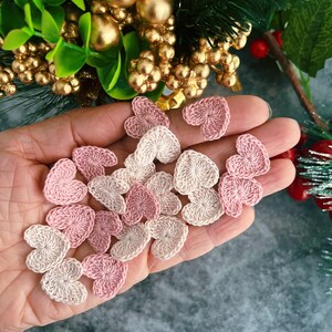 May include: A collection of small, crocheted hearts in varying shades of pink and cream are held in a hand. The hearts are arranged in a cluster, with a textured appearance. The background includes gold ornaments and greenery.