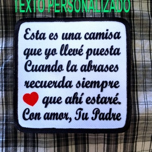 Op de afbeelding: Witte strijk-patch met zwarte tekst die "Esta es una camisa que yo llevé puesta Cuando la abrases recuerda siempre que ahí estaré. Con amor, Tu Padre" en een rood hart.