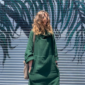 May include: A woman wearing a green, long-sleeved, collared dress with a relaxed fit. She is standing in front of a wall with a green and white abstract design.