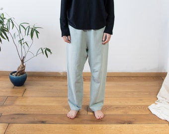 May include: A person wearing a black long-sleeved shirt and light blue pants. The person is standing on a wooden floor with a plant in a blue pot to the left.