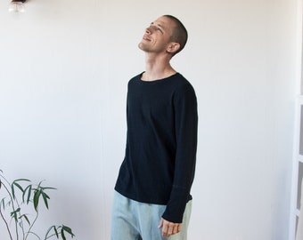 May include: A person wearing a black long-sleeve shirt and light blue pants. The pants are cuffed at the ankles. The person is standing on a wooden floor in front of a white wall. A small plant in a blue pot is to the left.