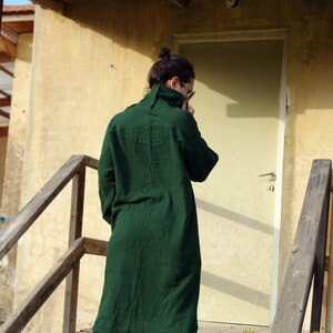 May include: A person wearing a long, dark green shirt and blue jeans stands on a set of concrete steps. The person is facing away from the camera and has their head down.