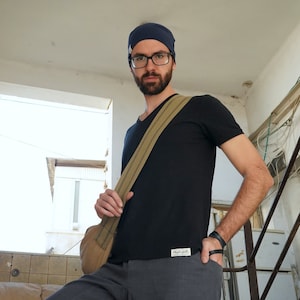 Könnte beinhalten: Ein Mann trägt ein schwarzes T-Shirt mit einem braunen Gurt, eine blaue Mütze und eine Brille. Er steht auf einer Treppe mit einer weißen Wand hinter ihm.