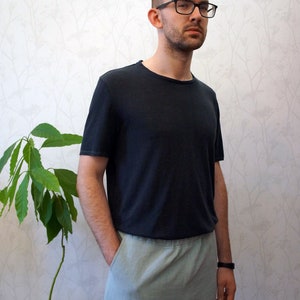 Könnte beinhalten: Ein Mann trägt ein schwarzes T-Shirt und graue Jogginghosen. Er steht vor einer weißen Wand mit einer grünen Pflanze in einem Topf.