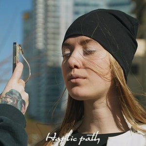 Könnte beinhalten: Eine Frau mit langen blonden Haaren, die eine schwarze Mütze und eine weiße Weste trägt, schaut auf ein Smartphone, das von einer Person mit einem Tattoo auf dem Arm gehalten wird. Der Text "Haptic path" ist auf der Weste sichtbar.