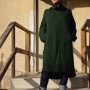 May include: A person wearing a dark green long-sleeved tunic with a high neck and pockets. They are wearing blue jeans with the cuffs rolled up and dark green slip-on shoes. The person is standing on a set of concrete steps.