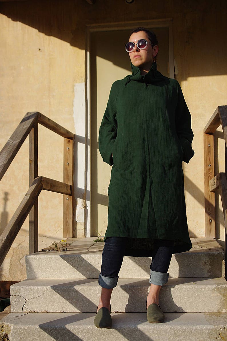 May include: A person wearing a dark green long-sleeved tunic with a high neck and pockets. They are wearing blue jeans with the cuffs rolled up and dark green slip-on shoes. The person is standing on a set of concrete steps.