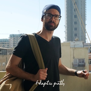 Könnte beinhalten: Ein Mann trägt ein schwarzes T-Shirt, eine blaue Mütze und eine Brille und trägt eine braune Canvas-Tasche. Er steht auf einem Balkon mit Blick auf die Stadt im Hintergrund.