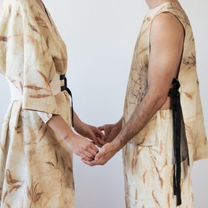 Könnte beinhalten: Zwei passende beigefarbene Leinen-Outfits mit botanischem Druck. Eines ist ein Kimono-Kleid mit weißem Gürtel und schwarzer Krawatte, das andere ein ärmelloses Top und Shorts mit schwarzen Krawatten. Die Outfits sind aus hellem Stoff gefertigt.