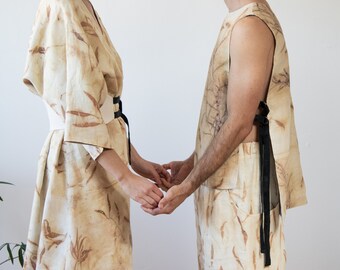 May include: Matching beige linen outfits with a botanical print. One outfit is a kimono-style dress with a white belt and black tie, the other is a sleeveless top and shorts with black ties. The outfits are made of a light-colored fabric.