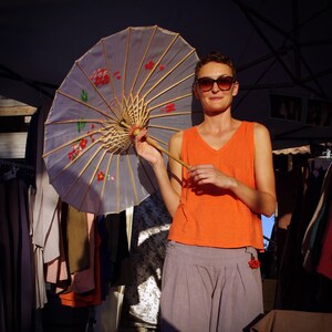 Könnte beinhalten: Eine Frau mit Sonnenbrille und einem leuchtend orangenen Tanktop hält einen großen, bunten Papier-Sonnenschirm mit einem Holzgriff. Sie steht vor einem Kleiderständer.
