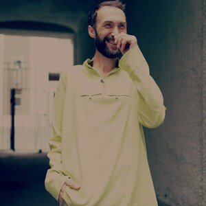 May include: A man with a beard is laughing and covering his mouth with his hand. He is wearing a light yellow long-sleeved shirt and khaki shorts.
