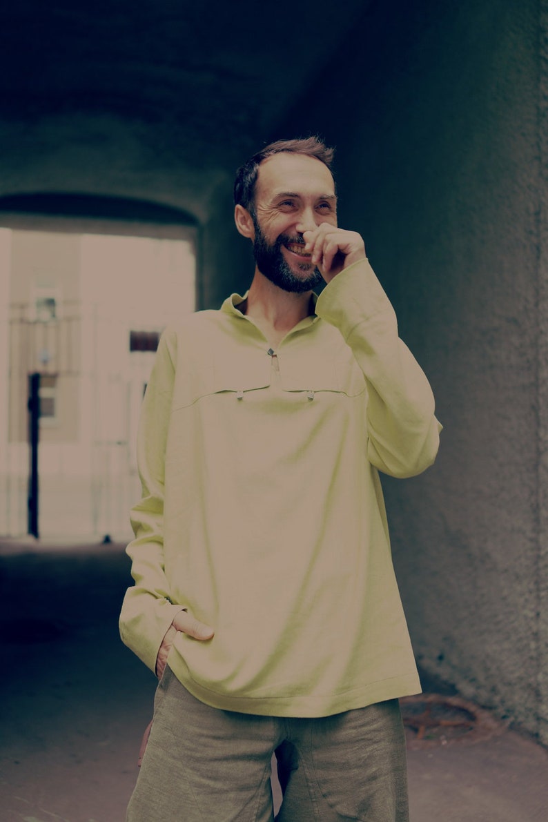 May include: A man with a beard is laughing and covering his mouth with his hand. He is wearing a light yellow long-sleeved shirt and khaki shorts.