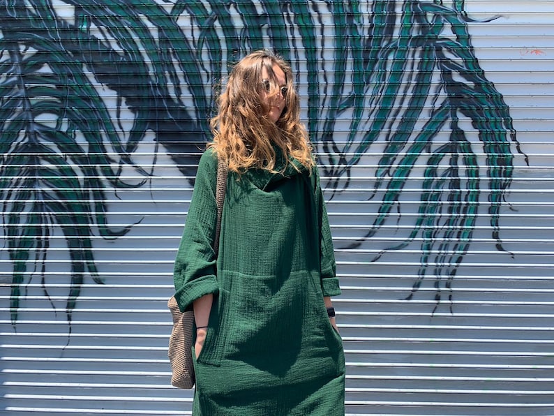 May include: A woman wearing a green, long-sleeved, collared dress with a relaxed fit. She is standing in front of a wall with a green and white abstract design.