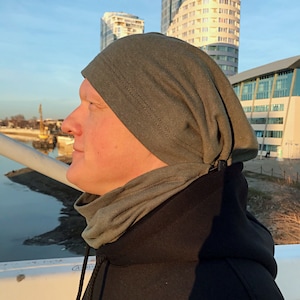 Könnte beinhalten: Eine Person trägt eine grüne Mütze und einen schwarzen Kapuzenpullover. Die Person steht vor einem Gebäude mit einer Glasfassade.